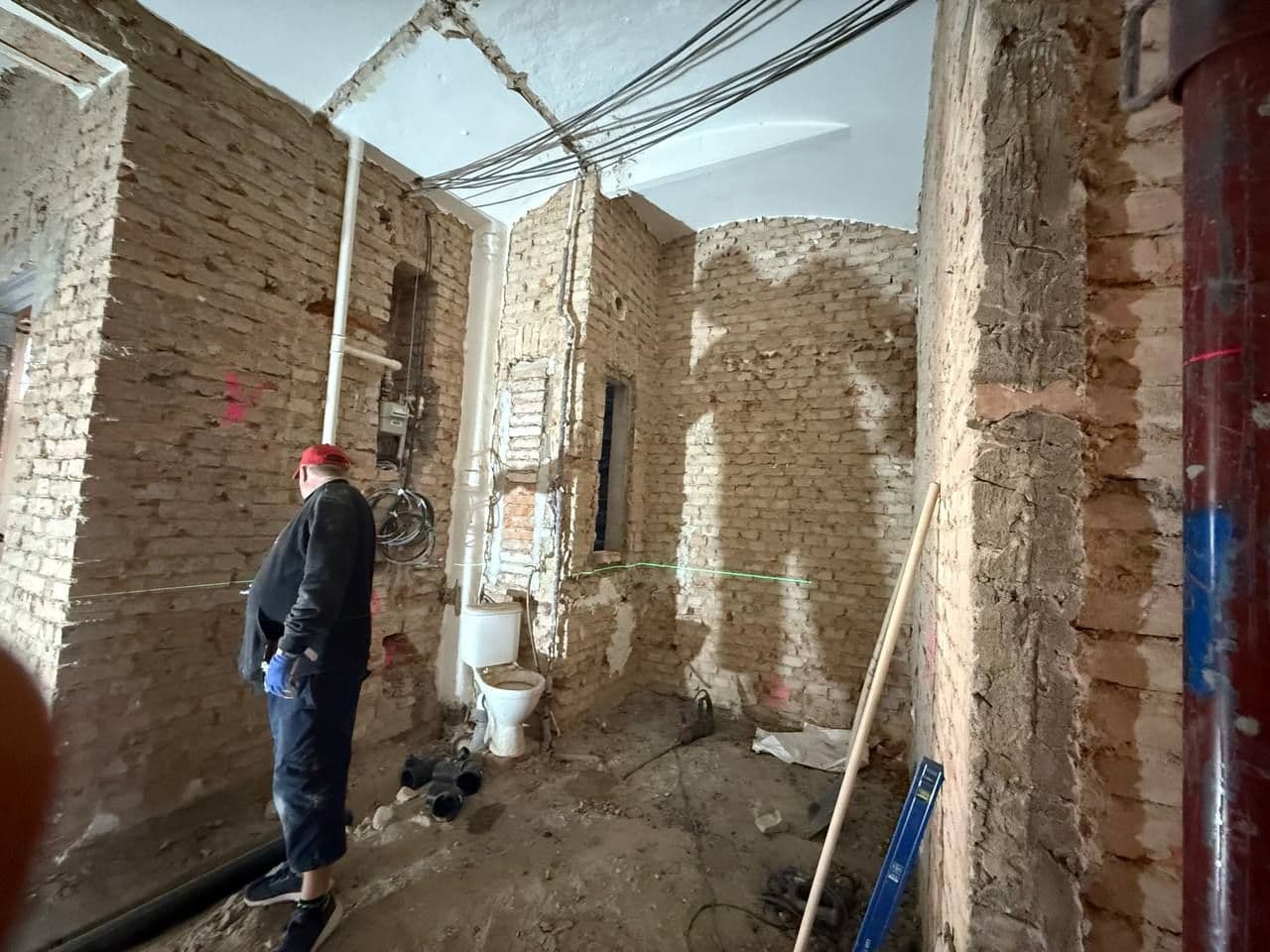 Pracovník v rozbourané místnosti s odkrytým stropem a vedením instalací, na podlaze suť.