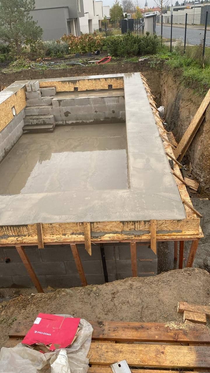 Hotová betonová vana bazénu se zabedněnou obrubou po obvodu, v pozadí zahrada a nová zástavba.